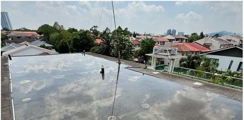 Standing water reflecting the sky across an old flat roof, houses and trees behind.