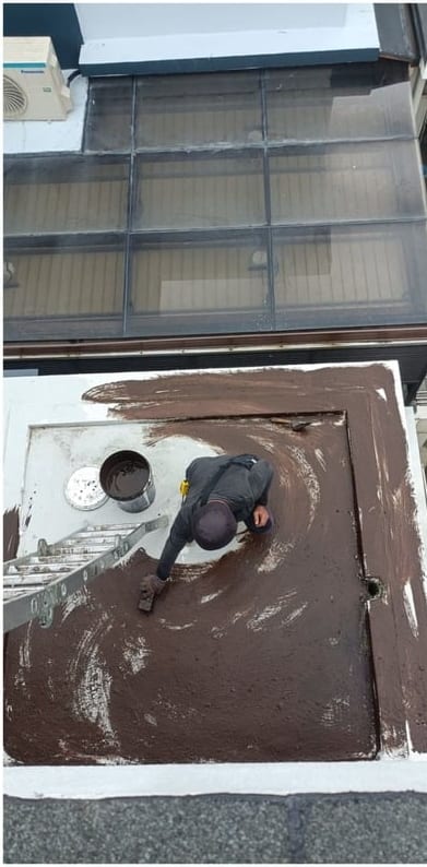 A worker kneeling on a small slab above a glass canopy, the surface partly covered in a brown coating.
