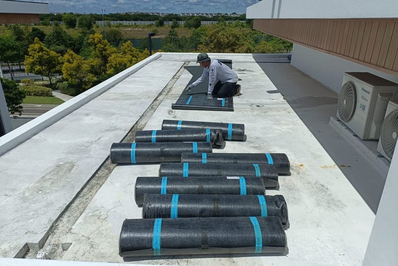 Rolls of membrane laid out across a white-parapet flat roof, a worker at the far end.
